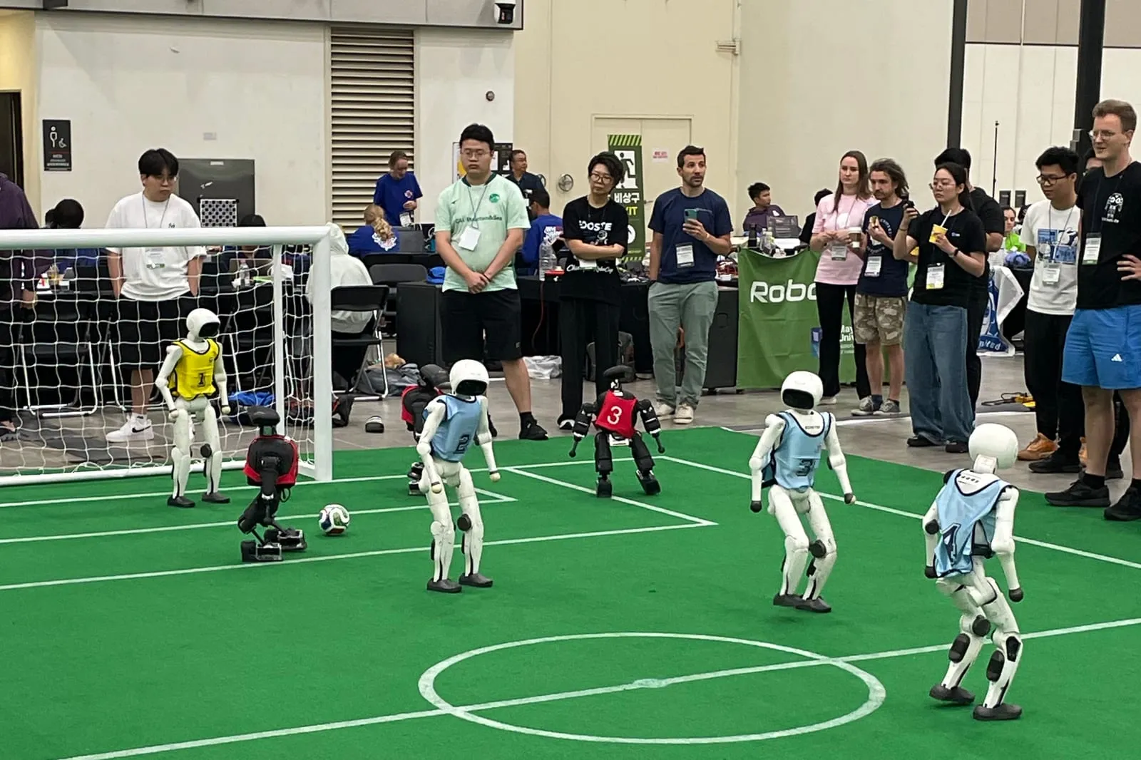 Humanoid robots in blue and red kit around the ball on a green pitch at a RoboCup match